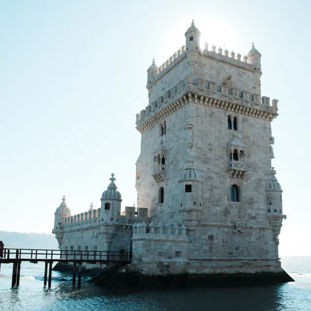 Belem Tower rises from the water with a wooden pier extending toward it, bright daylight illuminating the white stone, calm blue sea around.