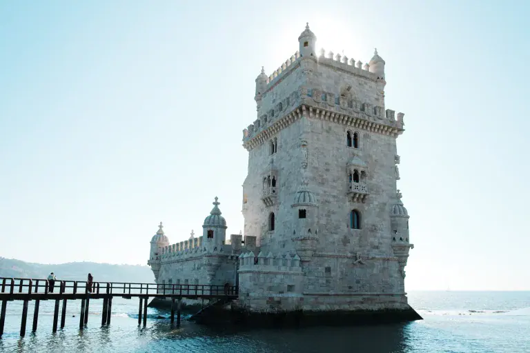 Belem Tower rises from the water with a wooden pier extending toward it, bright daylight illuminating the white stone, calm blue sea around.