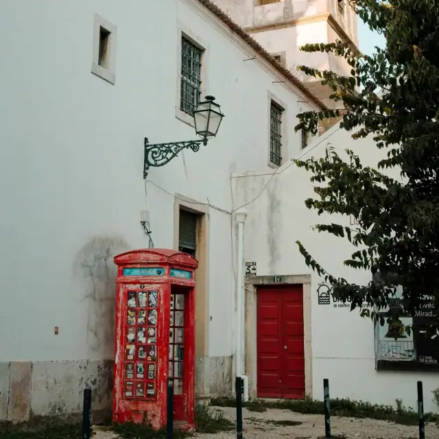 Old white building with red door, cobblestone street, and a classic red British-style phone booth near a small square.