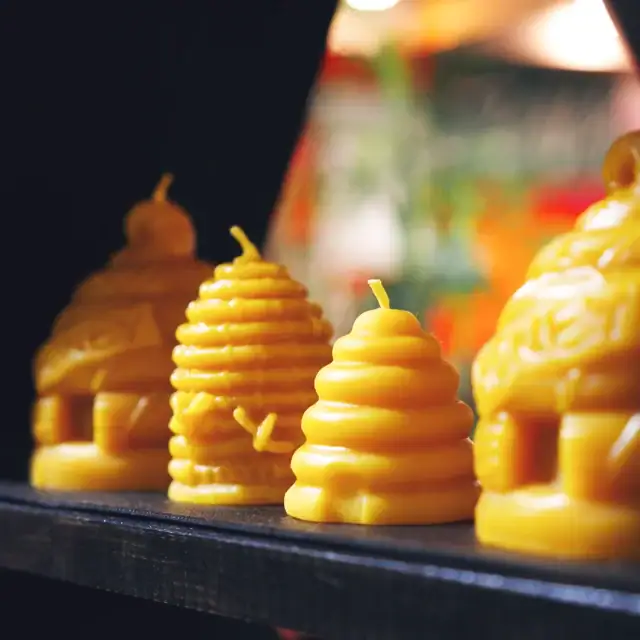 Close-up of yellow beeswax candles shaped like beehives and bees, arranged on a black shelf, with a blurred colorful background.