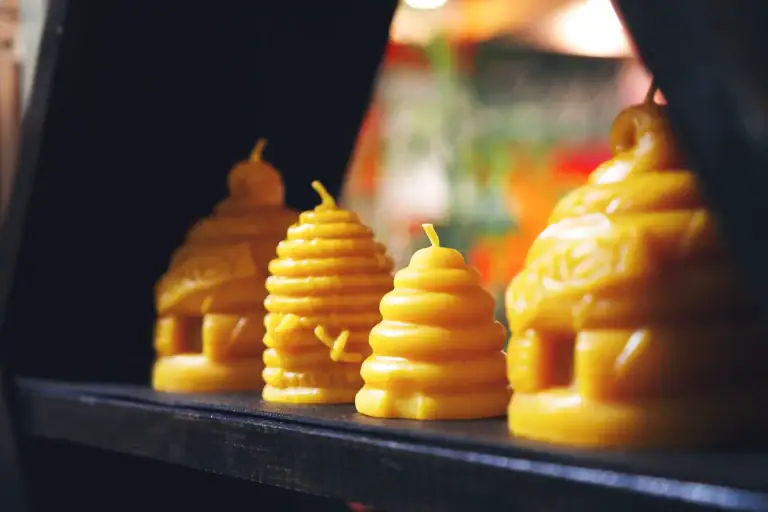 Close-up of yellow beeswax candles shaped like beehives and bees, arranged on a black shelf, with a blurred colorful background.
