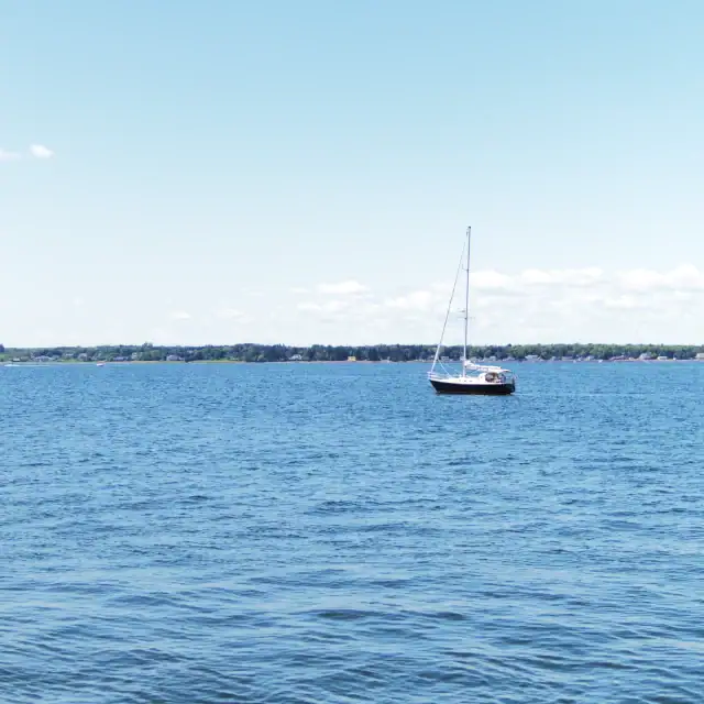 A sailboat floating peacefully on a calm blue lake under a clear sky, with a distant shoreline visible on the horizon.