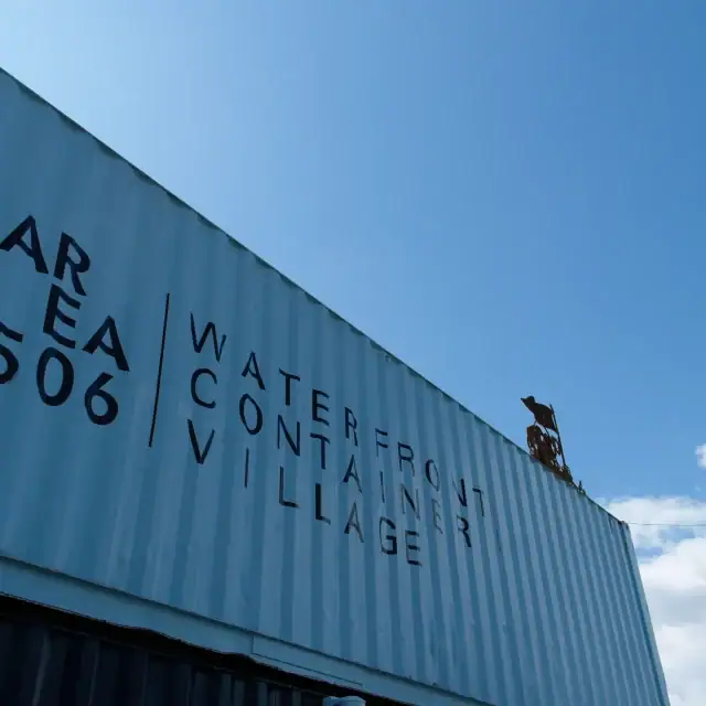 Close-up of a blue shipping container with the sign 'Waterfront Contain Village AR 506' in black letters, set against a bright blue sky.