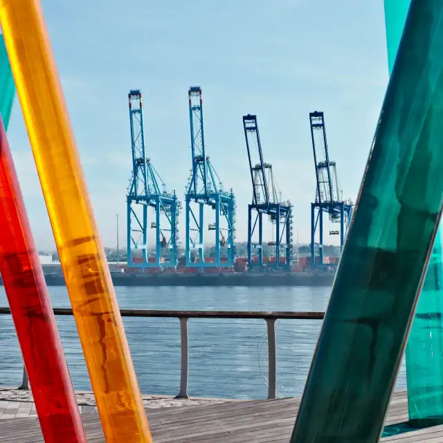 Vivid glass sculptures in red, orange, yellow, and teal along a waterfront promenade with industrial cranes in the background.