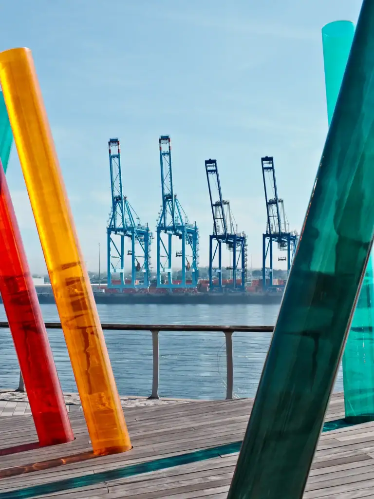 Vivid glass sculptures in red, orange, yellow, and teal along a waterfront promenade with industrial cranes in the background.