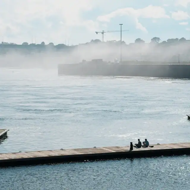 Two people sitting on a dock by the water with a misty river and industrial background, capturing a peaceful moment by the waterfront.