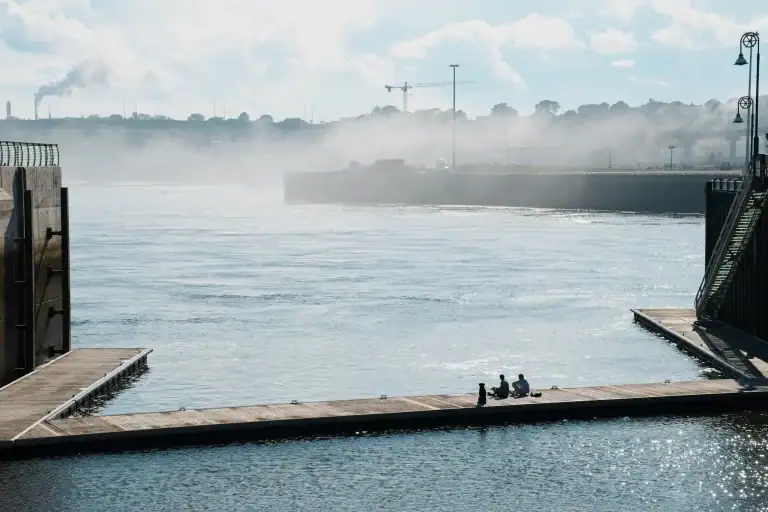 Two people sitting on a dock by the water with a misty river and industrial background, capturing a peaceful moment by the waterfront.