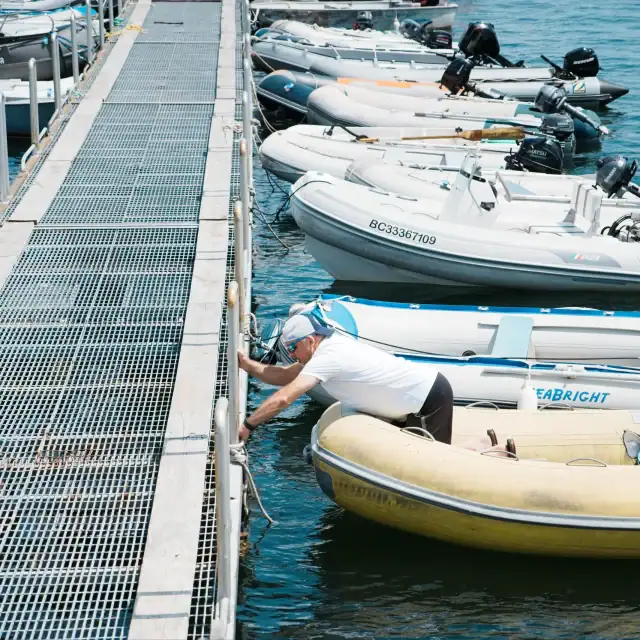 People preparing boats at a marina with a metal dock and various small boats, including inflatable and motorboats, on a sunny day.