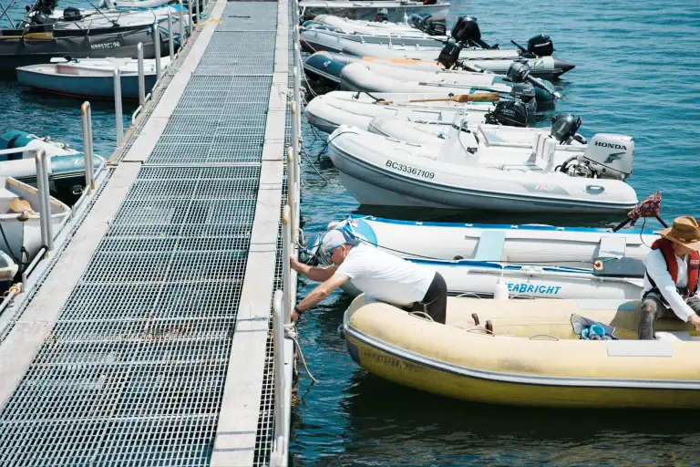 People preparing boats at a marina with a metal dock and various small boats, including inflatable and motorboats, on a sunny day.