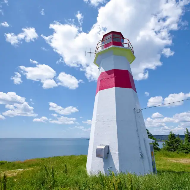 Colorful lighthouse with red and white stripes standing on grassy land under a bright blue sky with fluffy clouds.