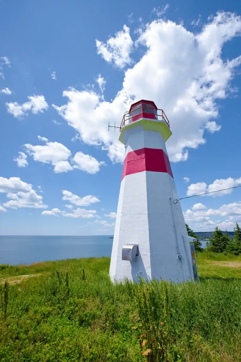 Colorful lighthouse with red and white stripes standing on grassy land under a bright blue sky with fluffy clouds.