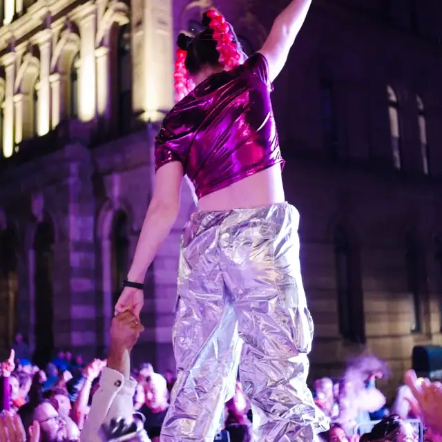 A performer in shiny silver pants and a purple top dances on stage during a nighttime event, with a crowd watching and a historic building illuminated in the background.