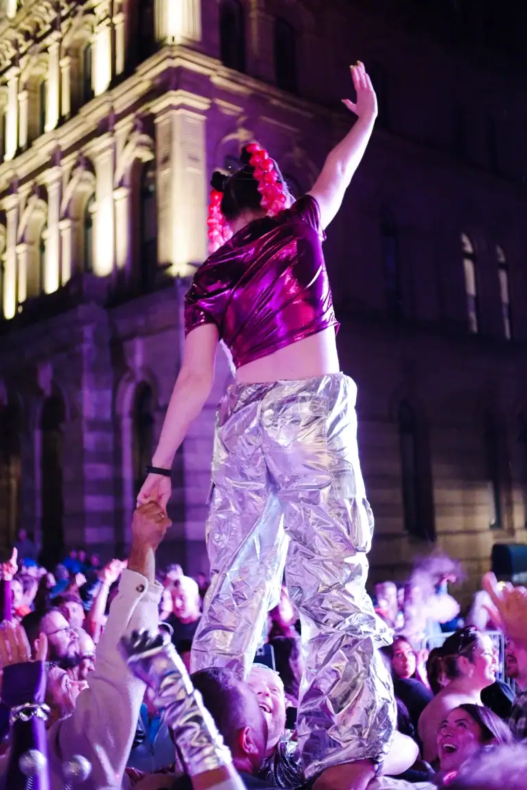 A performer in shiny silver pants and a purple top dances on stage during a nighttime event, with a crowd watching and a historic building illuminated in the background.