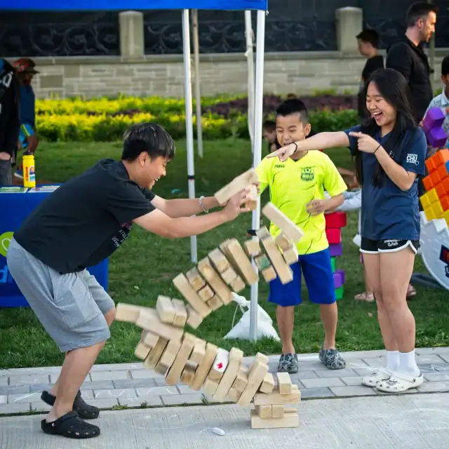 Children and adults enjoying a giant Jenga game outdoors at a community event with tents and greenery in the background.