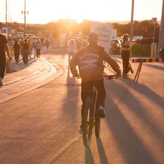 A cyclist riding a bike during sunset in East County, with people walking and cars in the background, capturing a lively outdoor scene.