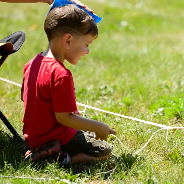 Young boy in a red shirt kneeling on grass, playing with a string outdoors, with an adult gently placing a blue hat on his head.