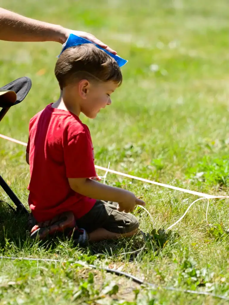 Young boy in a red shirt kneeling on grass, playing with a string outdoors, with an adult gently placing a blue hat on his head.
