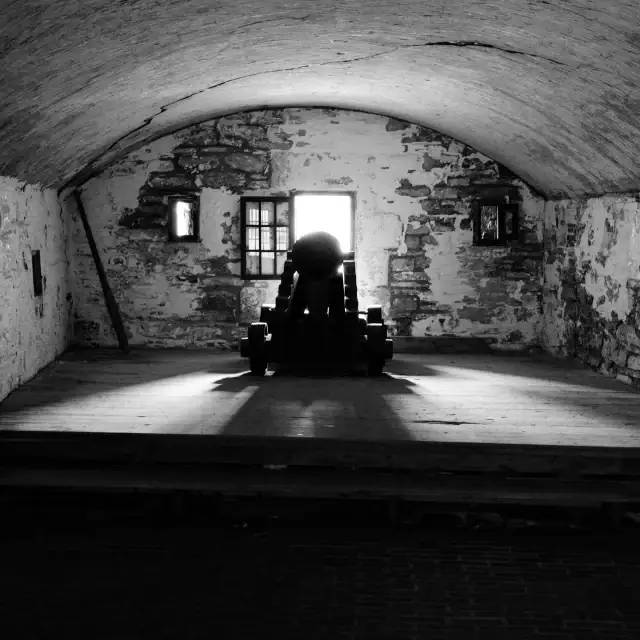 Black and white photo of a cannon inside an ancient stone fortress with a small window behind it, creating a silhouette effect.