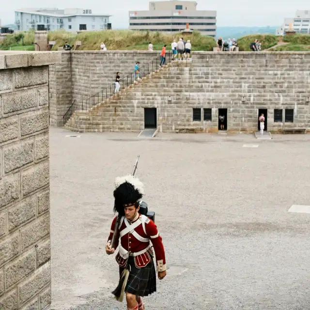 A person dressed in traditional Scottish Highland dress walking through a historic fortress with stone walls and stairs, with modern buildings in the background.