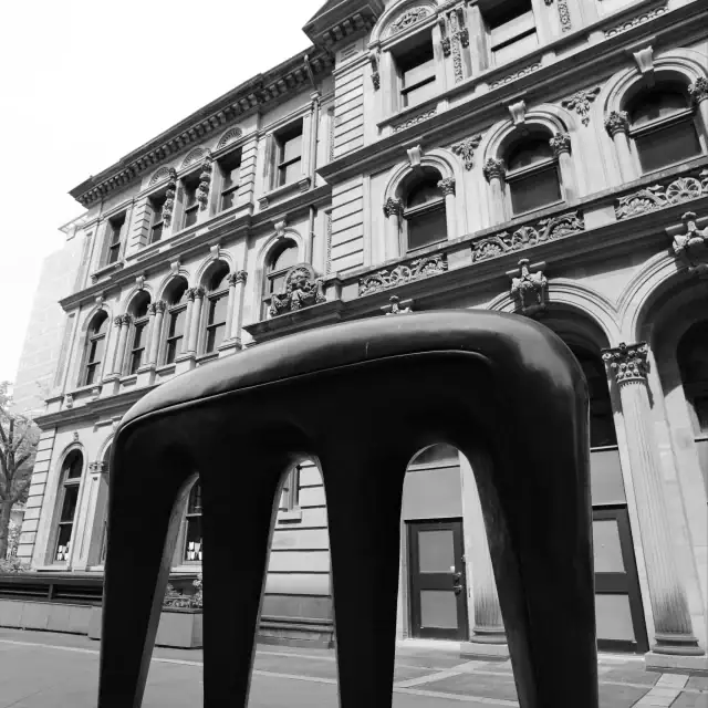 Black and white photo of an ornate historic building with a large, modern, black sculpture in the foreground on a city street.