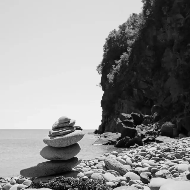 Balanced stack of smooth pebbles near a rocky shoreline by the sea, with a cliff in the background.