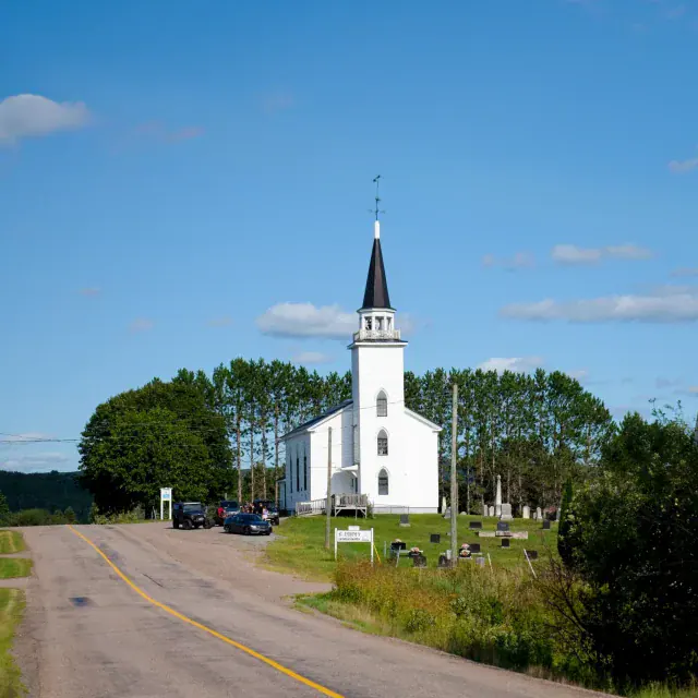A white church with a tall black steeple sits beside a winding rural road, surrounded by green trees and a clear blue sky.
