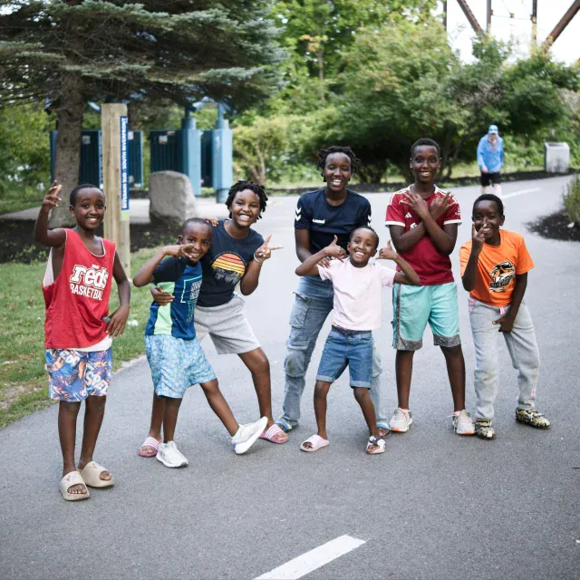 Group of smiling children posing together on a paved path in a park, some arms raised, bright daytime setting.