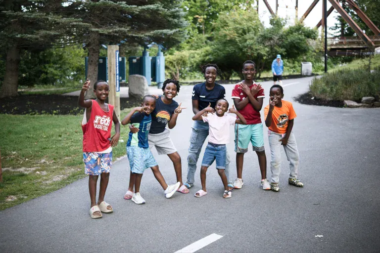Group of smiling children posing together on a paved path in a park, some arms raised, bright daytime setting.