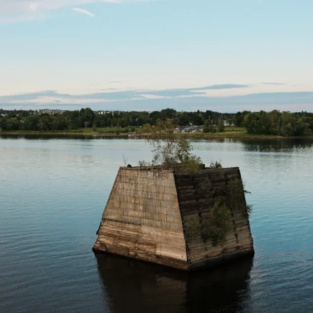 A solitary stone fort-like structure protrudes from calm river water, with greenery growing on its sides and a clear sky above.