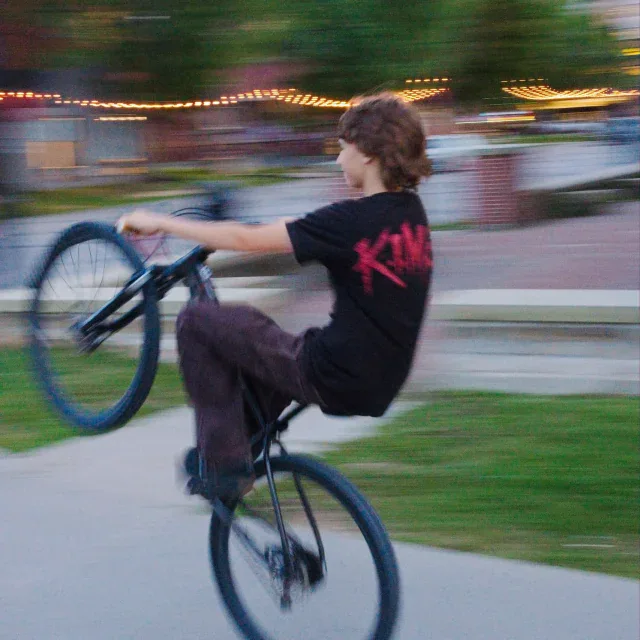 Young rider performing a wheelie on a BMX bike in a park with blurred motion and string lights in the background.