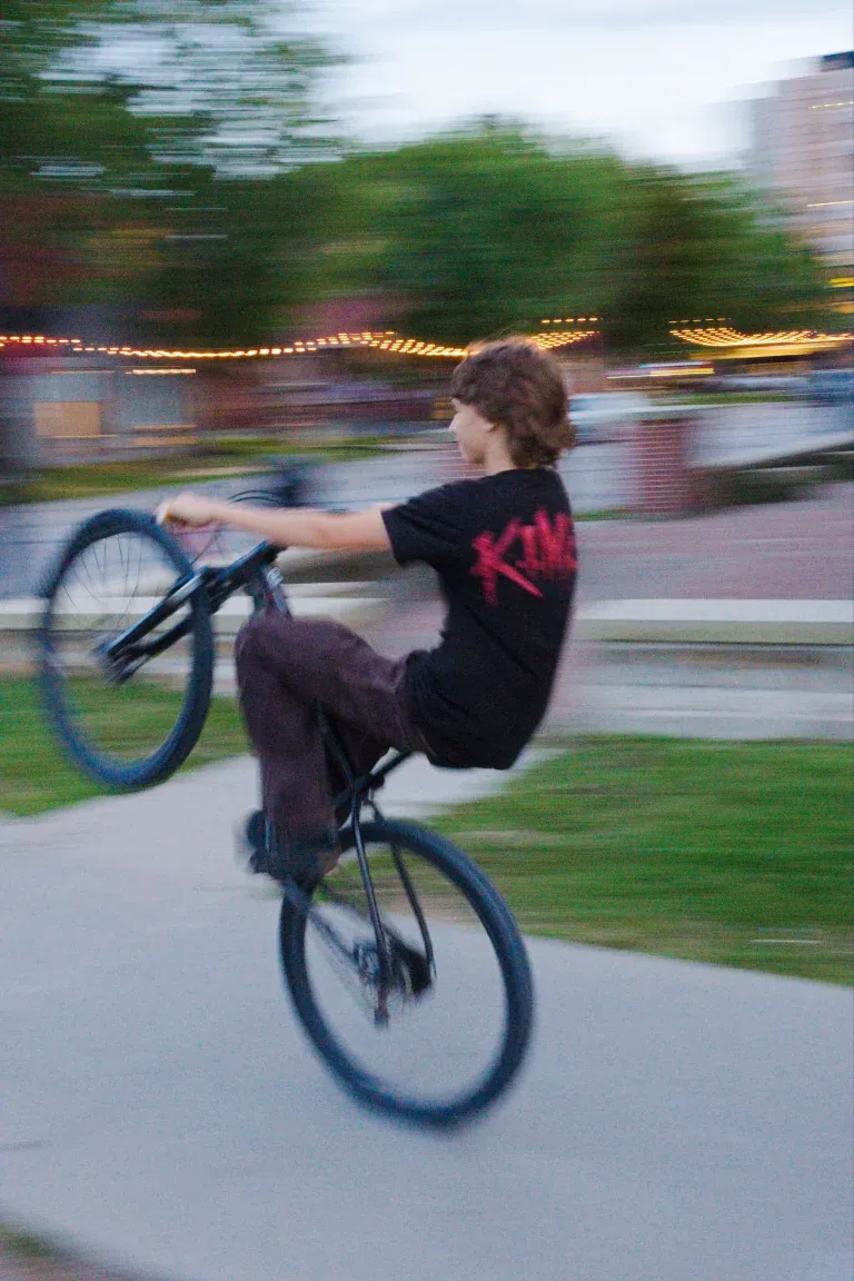 Young rider performing a wheelie on a BMX bike in a park with blurred motion and string lights in the background.
