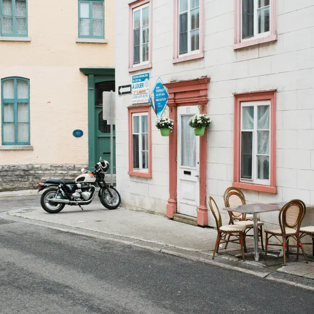 Quaint street scene with pastel-colored buildings, a vintage motorcycle parked by a curb, and outdoor café seating along the sidewalk.