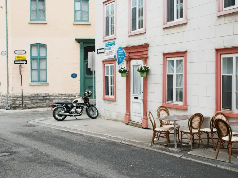 Quaint street scene with pastel-colored buildings, a vintage motorcycle parked by a curb, and outdoor café seating along the sidewalk.