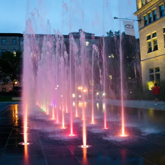 Night view of illuminated pink and purple water jets shooting upward from a modern plaza, with a historic building in the background.