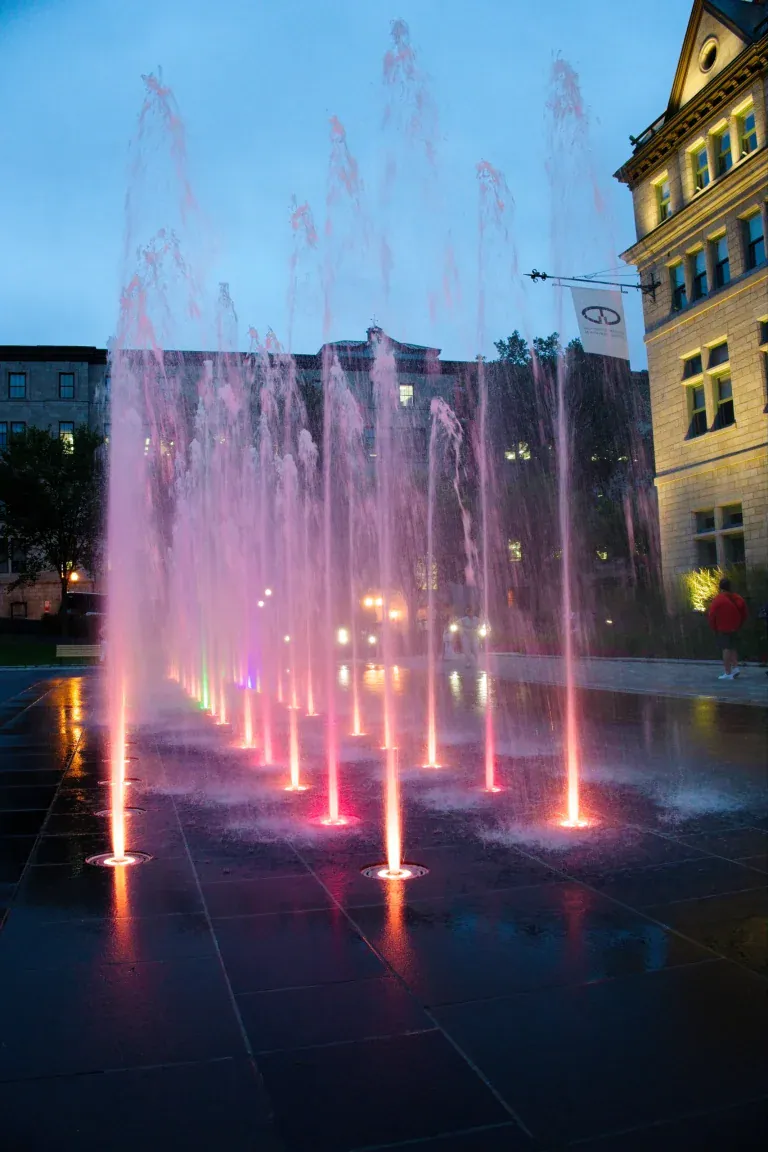 Night view of illuminated pink and purple water jets shooting upward from a modern plaza, with a historic building in the background.