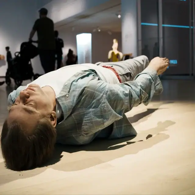 Person lying on the floor in a museum or gallery space, surrounded by onlookers and modern exhibit lighting.