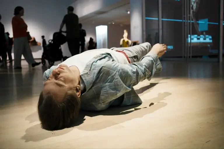 Person lying on the floor in a museum or gallery space, surrounded by onlookers and modern exhibit lighting.
