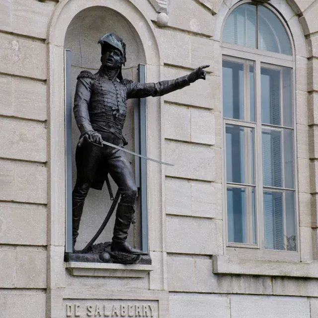 Bronze statue of a soldier in a historical uniform, standing in a niche on a stone building wall, with one arm extended and a sword sheathed.