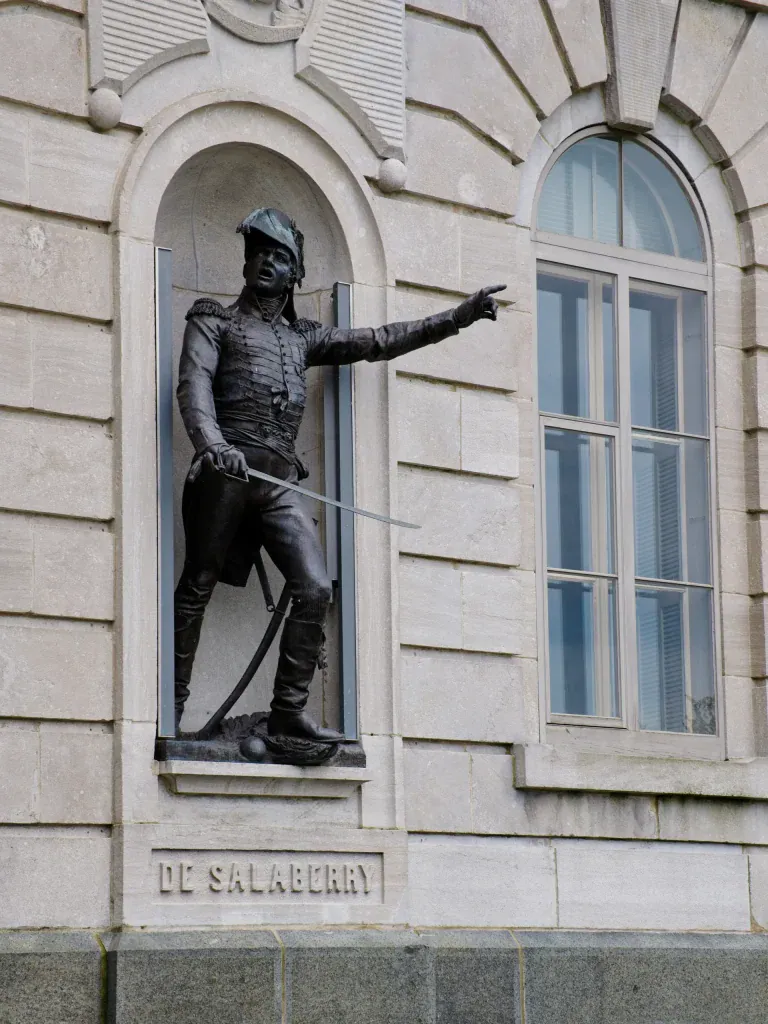Bronze statue of a soldier in a historical uniform, standing in a niche on a stone building wall, with one arm extended and a sword sheathed.