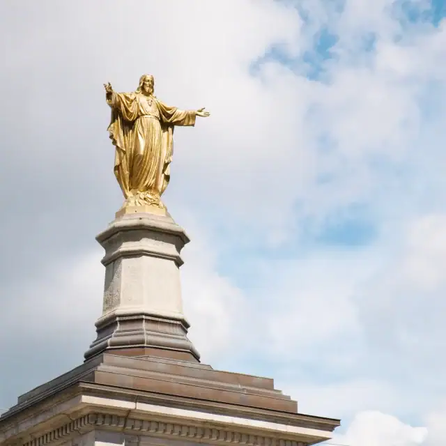 Golden statue of a robed figure with arms outstretched atop a stone pedestal against a blue sky with clouds.