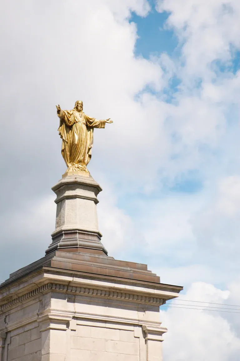 Golden statue of a robed figure with arms outstretched atop a stone pedestal against a blue sky with clouds.