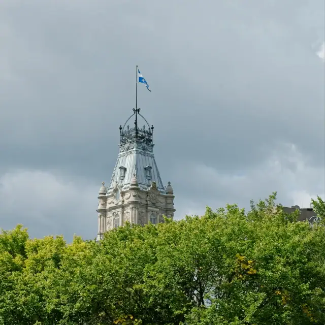Historic tower rising above lush green treetops, a flag fluttering atop the ornate spire against a cloudy sky.