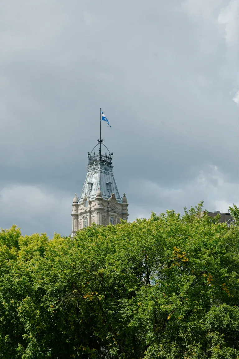 Historic tower rising above lush green treetops, a flag fluttering atop the ornate spire against a cloudy sky.
