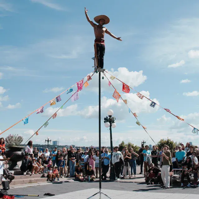 A shirtless street performer balances on a tall pole with arms raised, surrounded by a crowd of onlookers and colorful pennant banners, under a blue sky.