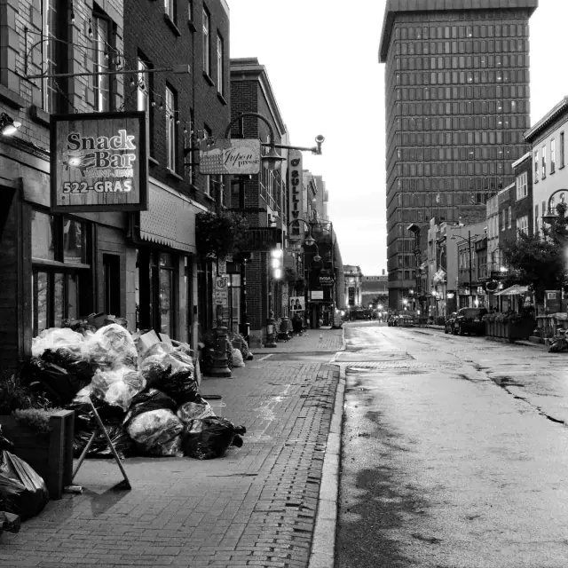 Black and white urban street image showing a row of buildings, a sidewalk piled with garbage bags, a bakery sign, and a distant tall building under an overcast sky.