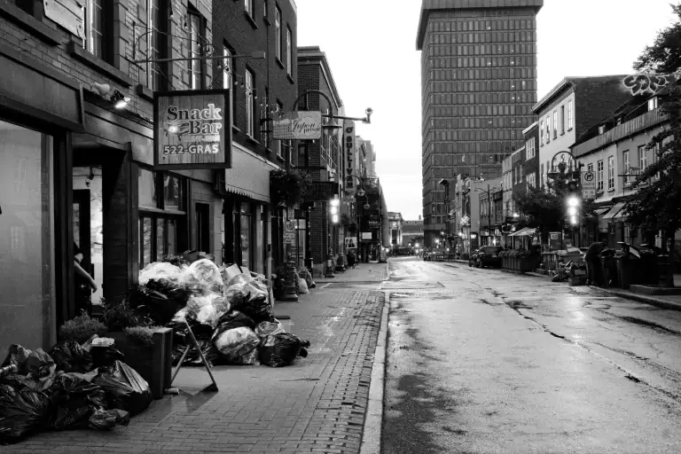 Black and white urban street image showing a row of buildings, a sidewalk piled with garbage bags, a bakery sign, and a distant tall building under an overcast sky.