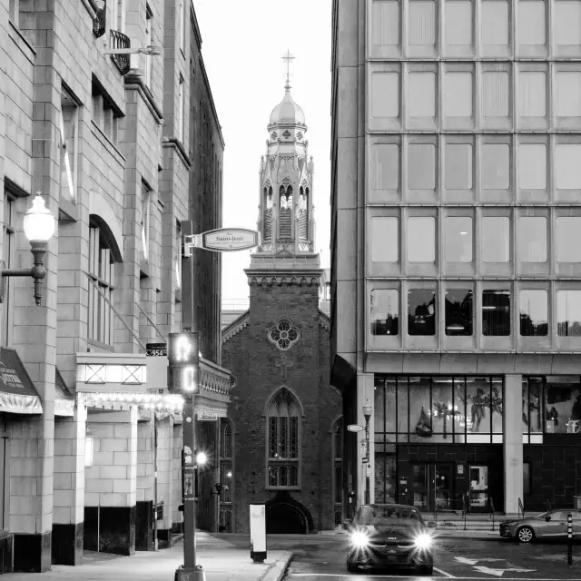 Black and white street scene featuring a church tower between tall buildings and a car on the road at dusk.
