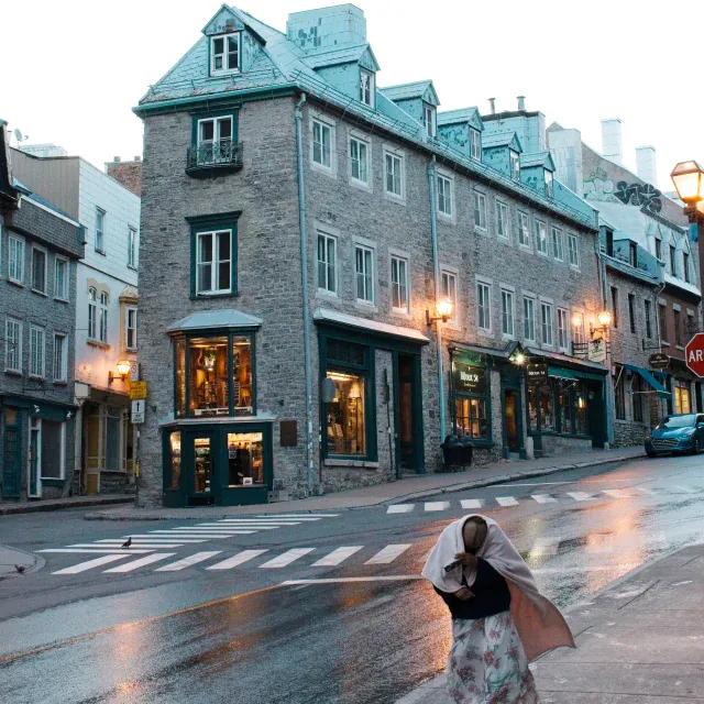 A lone person walking on a wet city street in front of brick townhouses with warm streetlights.