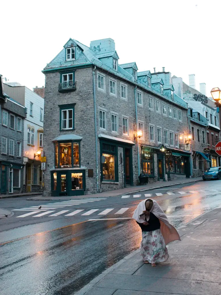 A lone person walking on a wet city street in front of brick townhouses with warm streetlights.