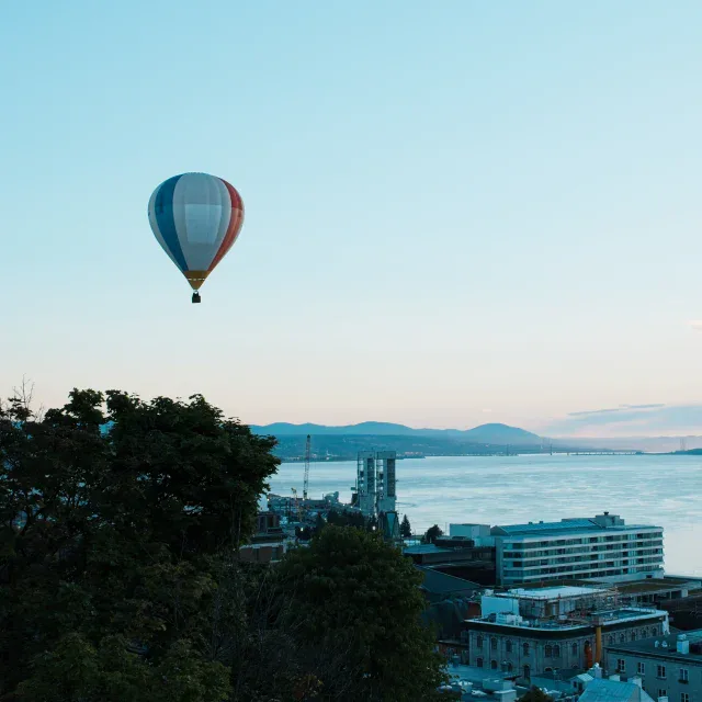 Blue and red hot air balloon floating above trees and riverfront buildings at sunset.